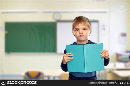 school, education and learning concept - displeased little boy reading book over classroom background. displeased little boy with book at school
