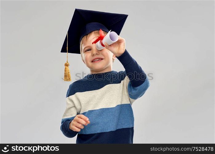 school, education and learning concept - happy little boy in bachelor hat or mortarboard looking through diploma scroll over grey background. little boy in mortarboard looking through diploma
