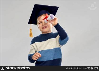 school, education and learning concept - happy little boy in bachelor hat or mortarboard looking through diploma scroll over grey background. little boy in mortarboard looking through diploma