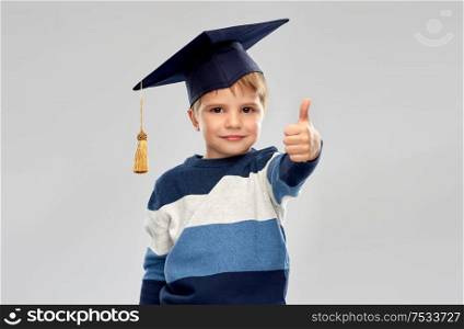 school, education and learning concept - happy little boy in bachelor hat or mortarboard showing thumbs up over grey background. little boy in mortarboard showing thumbs up