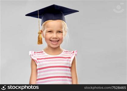 school, education and learning concept - happy little girl in bachelor hat or mortarboard over grey background. happy girl in bachelor hat or mortarboard