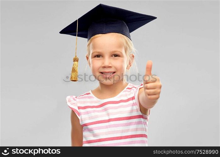 school, education and learning concept - happy little girl in bachelor hat or mortarboard showing thumbs up over grey background. little girl in mortarboard showing thumbs up