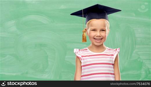 school, education and learning concept - happy little girl in bachelor hat or mortarboard over green chalkboard background. happy school girl in bachelor hat or mortarboard