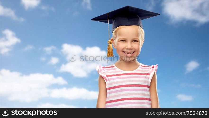school, education and learning concept - happy little girl in bachelor hat or mortarboard over blue sky and clouds background. happy girl in bachelor hat or mortarboard over sky