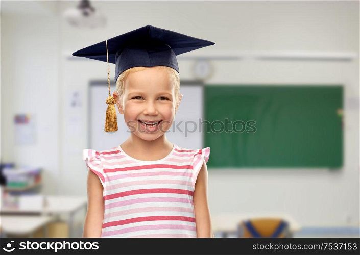 school, education and learning concept - happy little girl in bachelor hat or mortarboard over classroom background. happy girl in bachelor hat or mortarboard