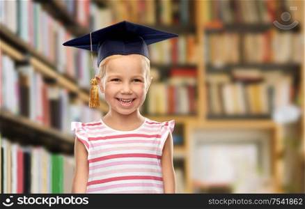 school, education and learning concept - happy little girl in bachelor hat or mortarboard over library book shelves background. girl in bachelor hat or mortarboard at library