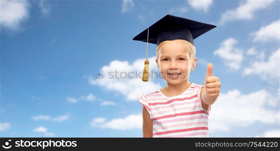 school, education and learning concept - happy little girl in bachelor hat or mortarboard showing thumbs up over blue sky and clouds background. little girl in mortarboard showing thumbs up