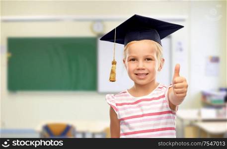 school, education and learning concept - happy little girl in bachelor hat or mortarboard showing thumbs up over classroom background. girl in mortarboard showing thumbs up at school
