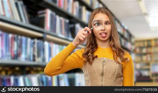 school, education and learning concept - happy teenage girl looking through magnifying glass and making silly faces over library background. teenage girl with magnifying glass at library