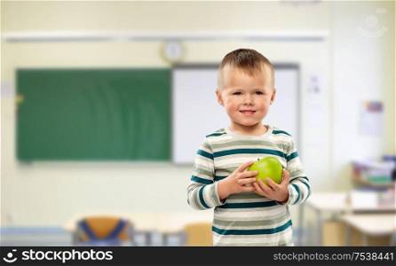 school, education and learning concept - portrait of smiling little boy holding green apple over classroom background. smiling boy holding green apple at school