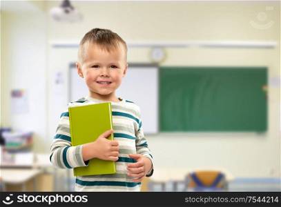school, education and learning concept - portrait of smiling little boy holding book over classroom background. portrait of smiling boy holding book at school