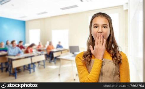 school, education and learning concept - scared or yawning teenage student girl closing her mouth by hand over classroom background. student girl closing her mouth by hand at school