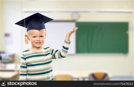 school, education and learning concept - smiling little boy in mortar board holding something in empty hand over classroom background. little boy in mortar board at school