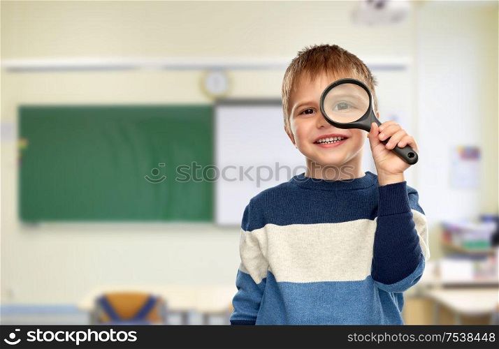 school, education and learning concept - smiling little boy looking through magnifying glass over classroom background. boy looking through magnifying glass at school