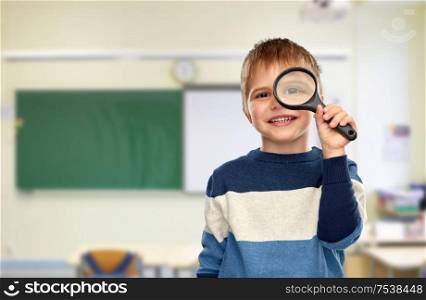 school, education and learning concept - smiling little boy looking through magnifying glass over classroom background. boy looking through magnifying glass at school