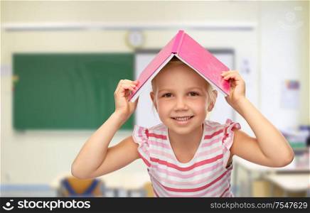 school, education and learning concept - smiling little girl with roof of book on top of her head over classroom background. little girl of book on top of her head at school