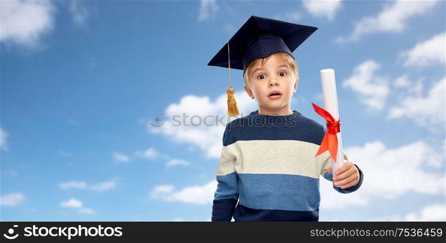 school, education and learning concept - surprised little boy in bachelor hat or mortarboard with diploma over blue sky and clouds background. little boy in mortarboard with diploma over sky