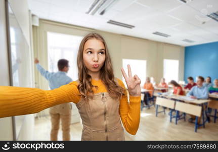 school, education and people concept - funny teenage student girl taking selfie and making faces over classroom background. teenage student girl taking selfie at school