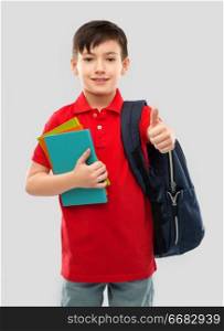 school, education and people concept - portrait of smiling little student boy in red polo t-shirt in glasses with books and bag showing thumbs up over grey background. boy with books and school bag showing thumbs up