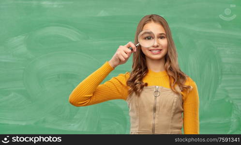 school, education and study concept - happy teenage student girl looking through magnifying glass over green chalk board background. student girl looking through magnifier at school