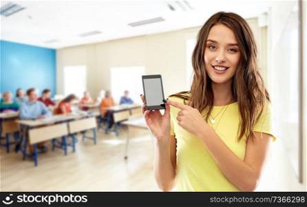 school, education and technology concept - smiling teenage student girl in yellow t-shirt pointing finger to smartphone blank screen over classroom background. teenage student girl holding smartphone at school