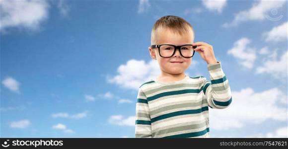 school, education and vision concept - portrait of smiling little boy in glasses over blue sky and clouds background. portrait of smiling boy in glasses over blue sky