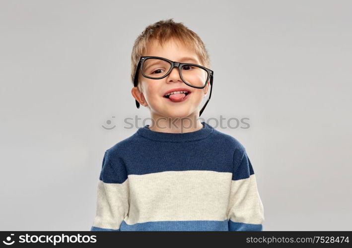 school, education and vision concept - portrait of smiling little boy with crookedly worn glasses showing tongue over grey background. portrait of little boy in glasses showing tongue