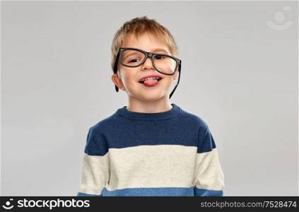 school, education and vision concept - portrait of smiling little boy with crookedly worn glasses showing tongue over grey background. portrait of little boy in glasses showing tongue