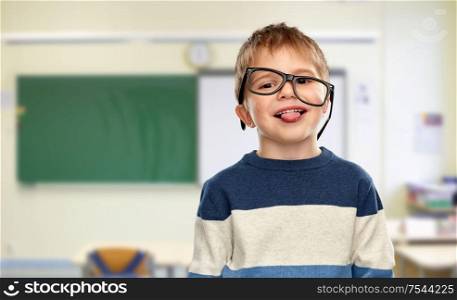 school, education and vision concept - portrait of smiling little boy with crookedly worn glasses showing tongue over classroom background. little boy in glasses showing tongue at school