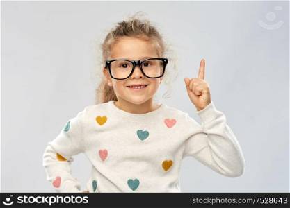 school, education and vision concept - portrait of smiling little girl in glasses pointing finger up over grey background. portrait of girl in glasses pointing finger up