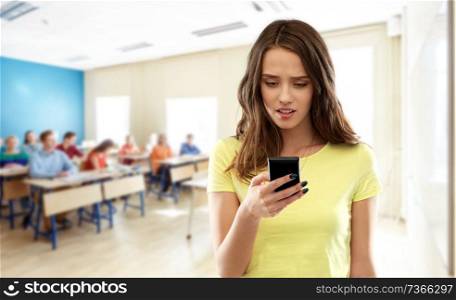 school, education, cyberbullying and technology concept - teenage student girl in blank yellow t-shirt using smartphone over classroom background. teenage student girl using smartphone at school