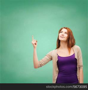 school, education, gesture and people concept - smiling teenage girl in casual clothes pointing finger up over green board background