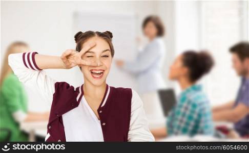 school, education, people, gesture and teens concept - happy smiling pretty teenage student girl showing peace sign and winking over classroom background with teacher and classmates