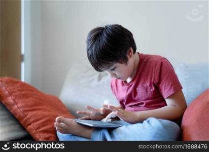 School kid sitting on sofa watching cartoon on tablet, Cute young boy playing game online on digital pad, Cinematic indoor portrait Child having fun and relaxing on his own in study room, Children with technology life style