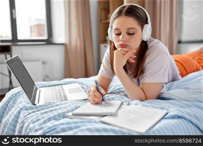 school, online education and e-learning concept - bored teenage student girl in headphones with laptop computer writing to notebook lying on bed at home. student girl in headphones learning at home