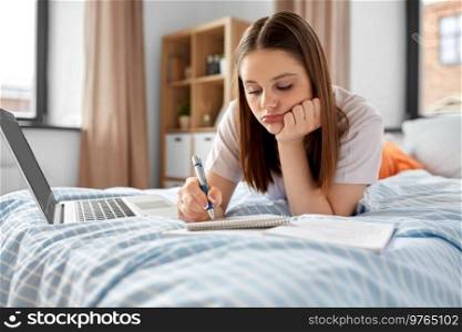 school, online education and e-learning concept - bored teenage student girl with laptop computer writing to notebook lying on bed at home. teenage student girl with laptop learning at home
