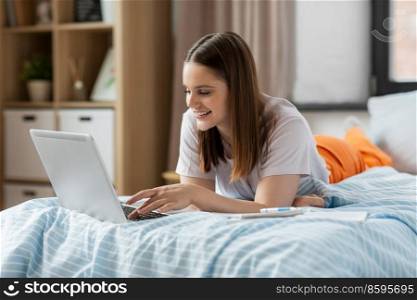 school, online education and e-learning concept - happy smiling teenage student girl with laptop computer writing to notebook lying on bed at home. teenage student girl with laptop learning at home