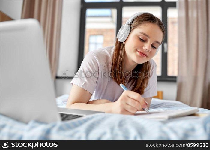 school, online education and e-learning concept - happy smiling teenage student girl in headphones with laptop computer writing to notebook lying on bed at home. happy student girl in headphones learning at home