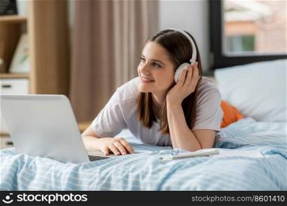 school, online education and e-learning concept - happy smiling teenage student girl in headphones with laptop computer lying on bed and listening to music at home. happy student girl in headphones learning at home