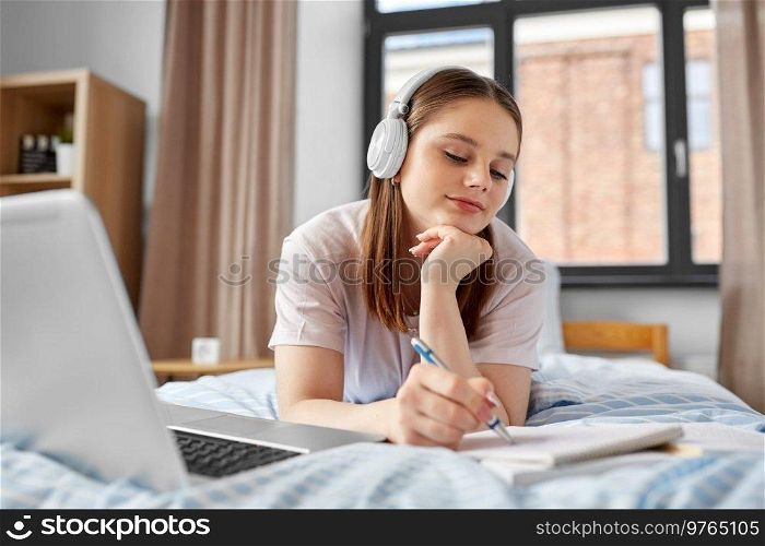 school, online education and e-learning concept - happy smiling teenage student girl in headphones with laptop computer writing to notebook lying on bed at home. happy student girl in headphones learning at home