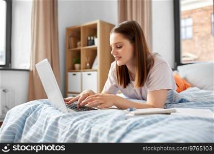 school, online education and e-learning concept - teenage student girl with laptop computer writing to notebook lying on bed at home. teenage student girl with laptop learning at home