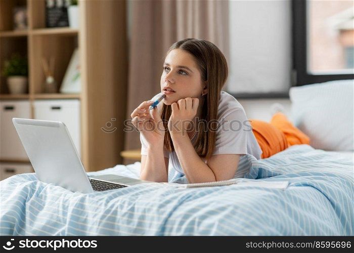 school, online education and e-learning concept - thinking or daydreaming teenage student girl with laptop computer writing to notebook lying on bed at home. teenage student girl writing to notebook at home