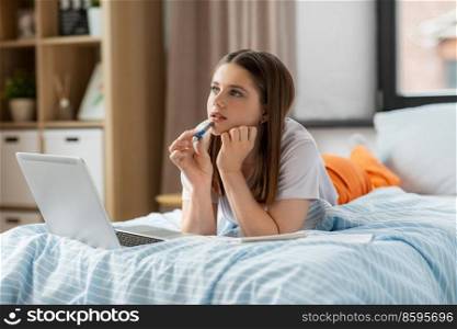 school, online education and e-learning concept - thinking or daydreaming teenage student girl with laptop computer writing to notebook lying on bed at home. teenage student girl writing to notebook at home