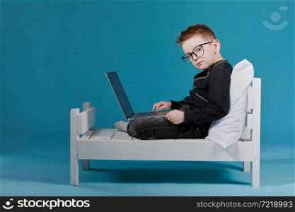 schoolboy in glasses using laptop sitting on bed. Child doing school homework and looking on camera. Online learning, distance lesson, education with online tutor at home isolation.. schoolboy in glasses using laptop sitting on bed. Child doing school homework and looking on camera. Online learning, distance lesson, education with online tutor at home isolation