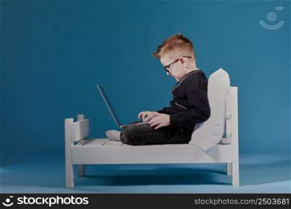 schoolboy in glasses using laptop sitting on bed. Child doing school homework. Online learning, distance lesson, education with online tutor at home isolation.. schoolboy in glasses using laptop sitting on bed. Child doing school homework. Online learning, distance lesson, education with online tutor at home isolation
