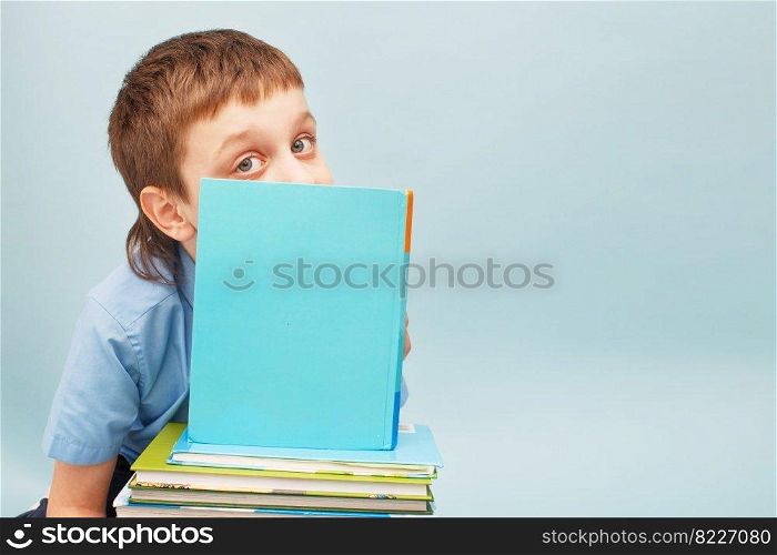 Schoolboy is sitting with a stack of books and reading and covers his face with a book isolated on a blue background. Schoolboy is sitting with a stack of books and reading and covers his face with a book isolated on blue background