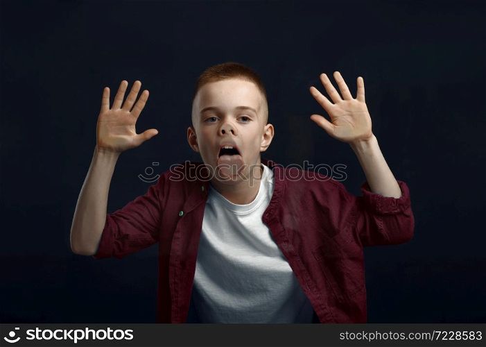 Schoolboy makes face leaning against the glass in studio. Happy childhood, children having fun, funny kid isolated on dark background, child emotion. Schoolboy makes face leaning against the glass