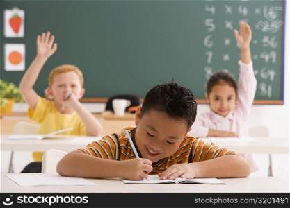Schoolboy writing on a notebook with a pencil and two students sitting in the background with their hands raised