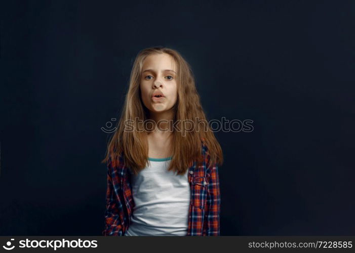Schoolgirl makes face leaning against the glass in studio. Happy childhood, children having fun, funny kid isolated on dark background, child emotion. Schoolgirl makes face leaning against the glass