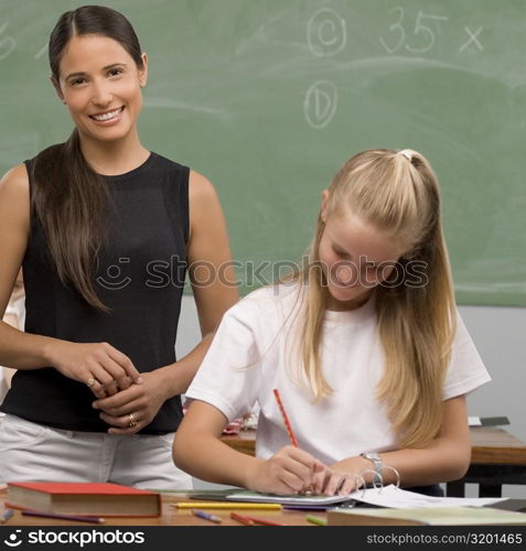 Schoolgirl writing on a textbook with her female teacher standing behind her and smiling